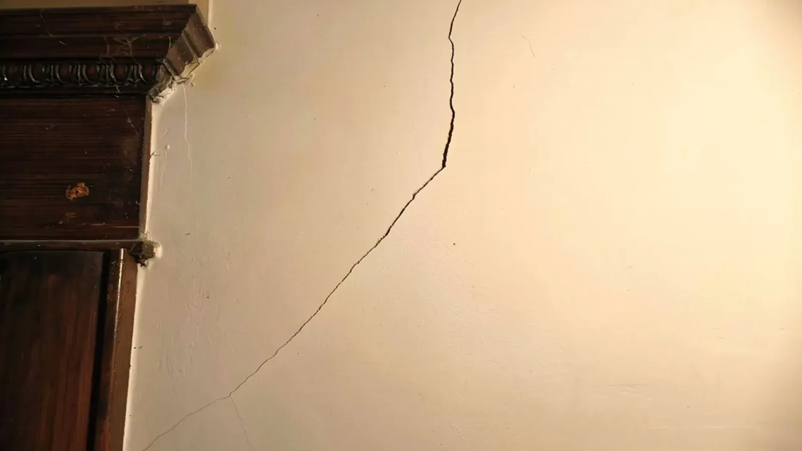 A fine hairline crack running diagonally across a painted interior wall from the corner of a dark-wood door casing in an older home