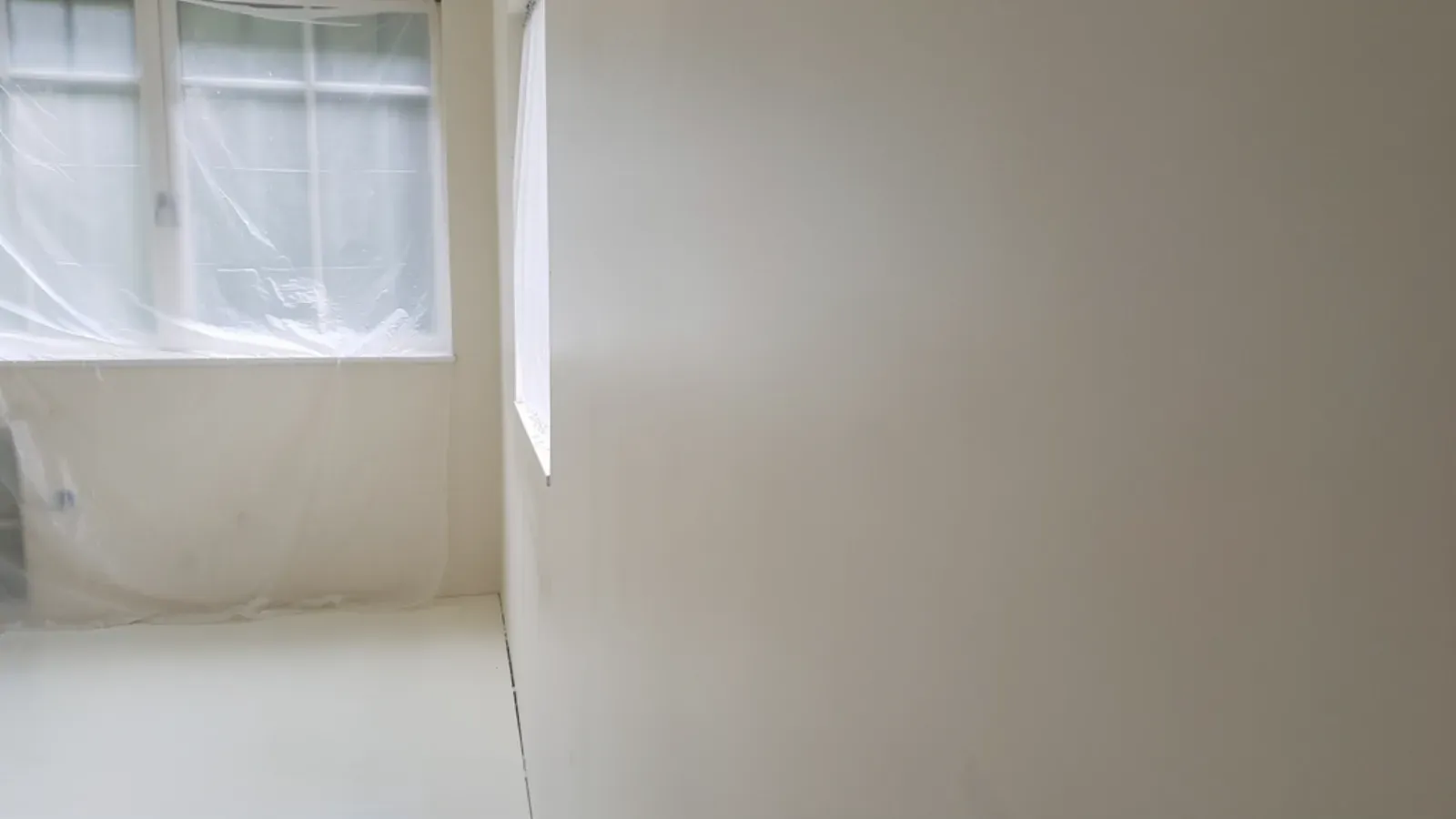 A freshly skim-coated interior wall running back from a plastic-masked window, the surface an even off-white with no visible seams